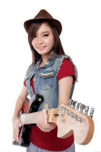 Pretty Asian guitarist girl in jeans jacket and fedora hat smiles at camera while holding her electric guitar, on white background